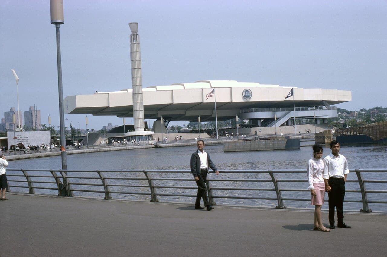 A colour photograph of a world's fair. A wide, low, white building shaped like a stack of offset slabs sits on supports beside a lagoon, with a tall slender tower rising through it. People walk along a railed promenade in the foreground.