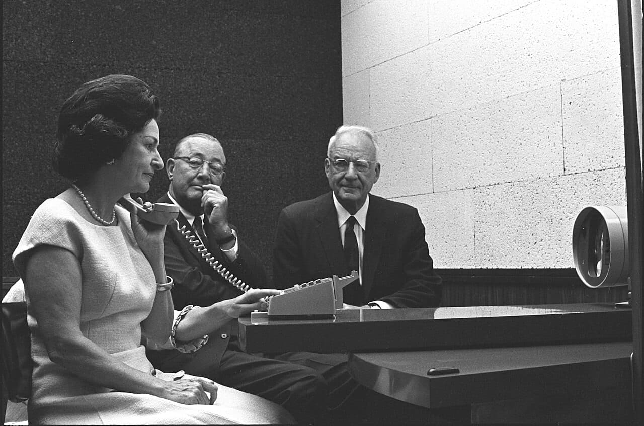 A black and white photograph. A seated woman in a pale sleeveless dress holds a small telephone handset to her ear and rests her other hand on a keypad on the desk in front of her. Two men in dark suits sit beside her. At the right edge of the frame stands a rounded, tapering set with a small screen and a lens.