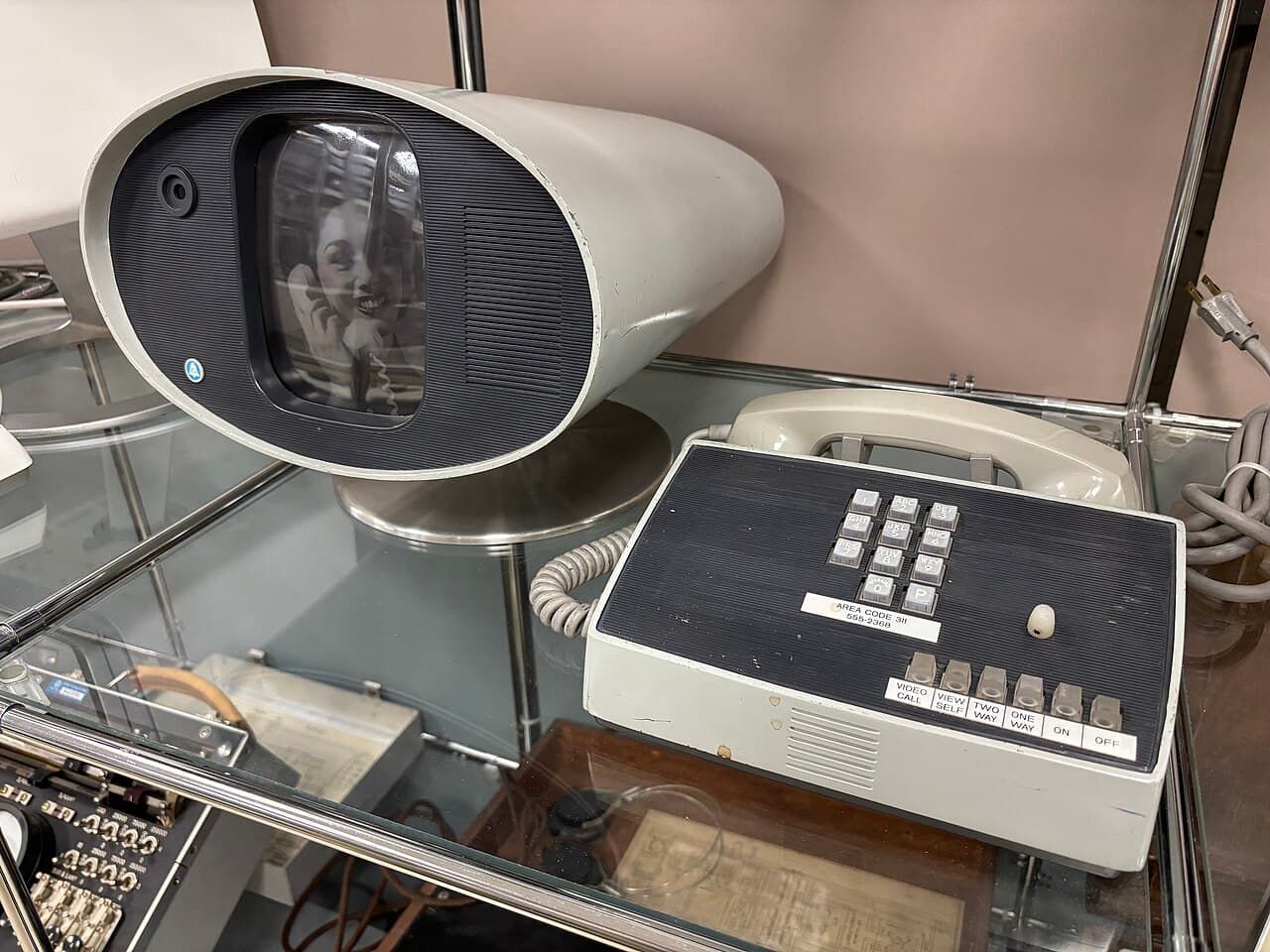 A colour photograph in a museum display case. A grey, tapering, oval-mouthed set stands on a chrome pedestal, holding a small screen and a lens; a face is visible on the screen. Beside it sits a cream and black keypad unit with buttons labelled VIDEO CALL, VIEW SELF, TWO WAY, ONE WAY, ON and OFF.