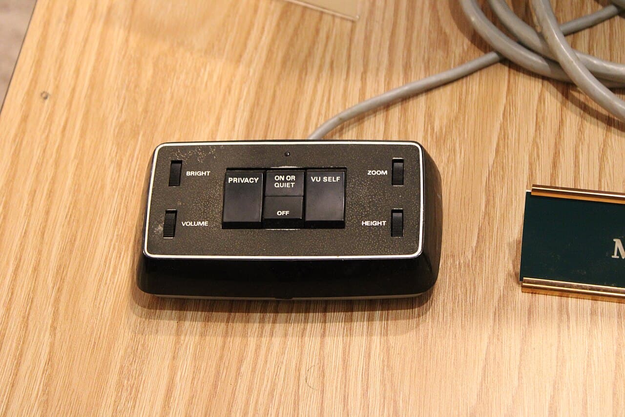 A colour photograph of a small black control pad on a wooden table. Four sliders at its corners are labelled BRIGHT, VOLUME, ZOOM and HEIGHT. Four buttons across the middle are labelled PRIVACY, ON OR QUIET, OFF and VU SELF.
