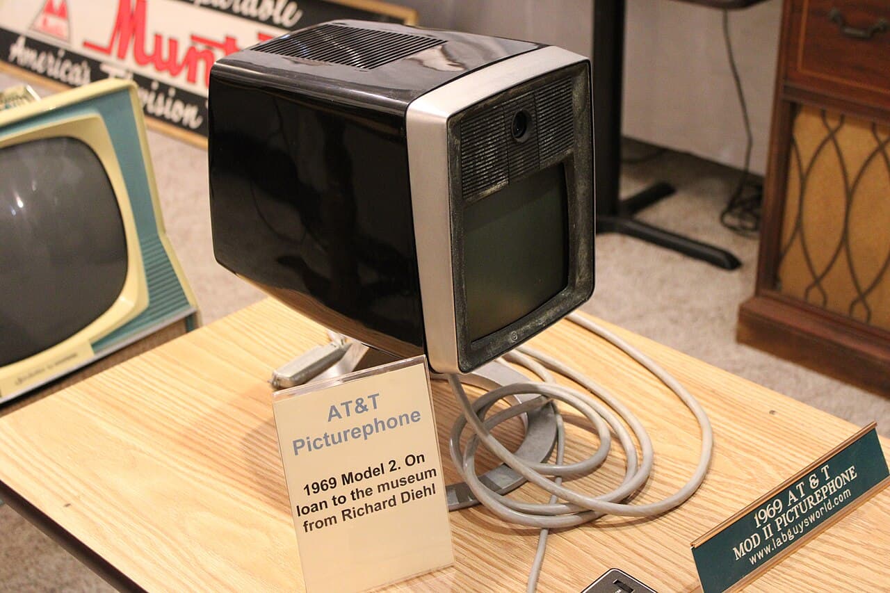 A colour photograph of a black and silver desktop unit on a wooden table. It has a small, roughly square dark screen, a lens set above the screen behind a grille, and a thick coiled grey cable. Two printed labels stand beside it.